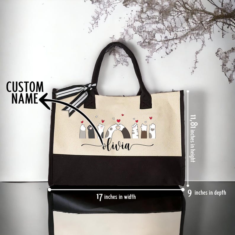 May include: A beige and brown canvas tote bag with black handles. The bag features a design of cats with hearts and the name "olivia" printed on the front. The bag measures 17 inches wide, 11.81 inches high, and 9 inches deep.