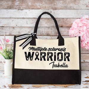 May include: A canvas tote bag with black handles and a black bottom panel. The bag features the text "Multiple sclerosis WARRIOR Isabella" with a black awareness ribbon graphic and a black and white striped bow.