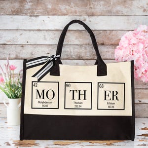 May include: A beige canvas tote bag with black handles and a black bottom. The bag has three squares with the periodic table elements for Molybdenum, Thorium, and Erbium, spelling out the word "MOTHER".