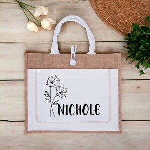 May include: A brown and white tote bag with a white pocket featuring a black line drawing of two flowers and the name "NICHOLE" in black text.