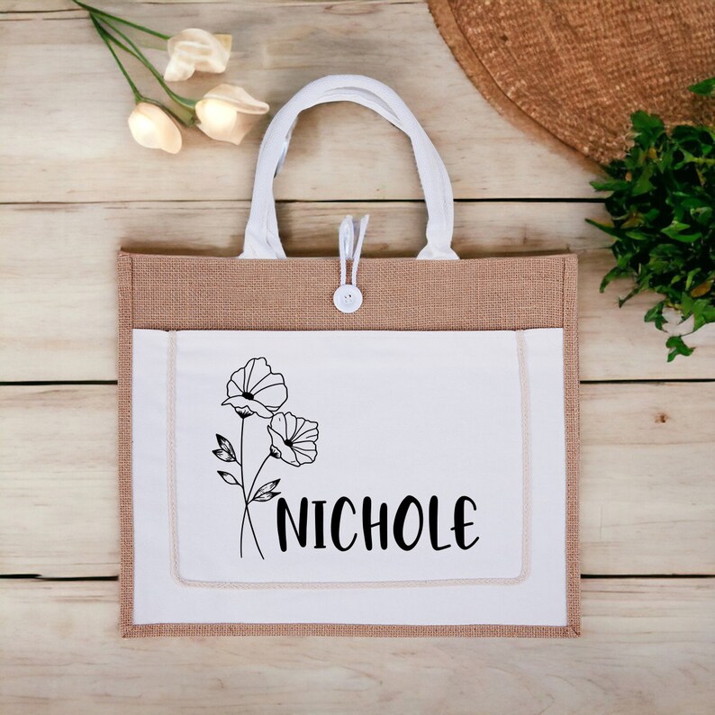 May include: A brown and white tote bag with a white pocket featuring a black line drawing of two flowers and the name "NICHOLE" in black text.