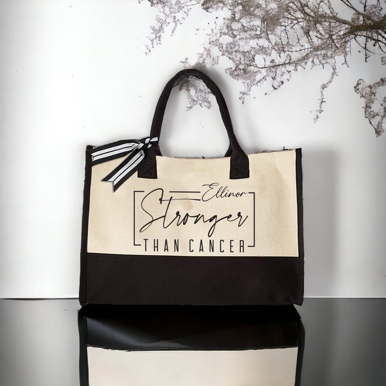 May include: A beige and brown canvas tote bag with black handles and a black and white striped ribbon. The bag has the text "Ellinor Stronger Than Cancer" printed on the front.