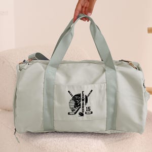 May include: A light green duffel bag with a black hockey helmet and crossed hockey sticks design. The text "EDWARD" and the number "15" are also embroidered on the bag.