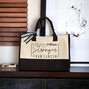 May include: A beige and black canvas tote bag with black handles and a black and white ribbon. The bag has the text "Valeria Stronger Than Cancer" printed on the front.