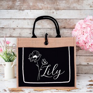 May include: A black and tan jute tote bag with a black fabric pocket featuring a white poppy flower design and the name "Lily" in white script.