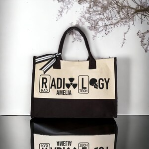 May include: A black and white canvas tote bag with the word "RADIOLOGY" printed on the front. The word is broken up into three sections: "RAD", "AMELIA", and "TECH". Each section is surrounded by a black rectangle. A skull and crossbones symbol is printed to the right of the word "TECH".