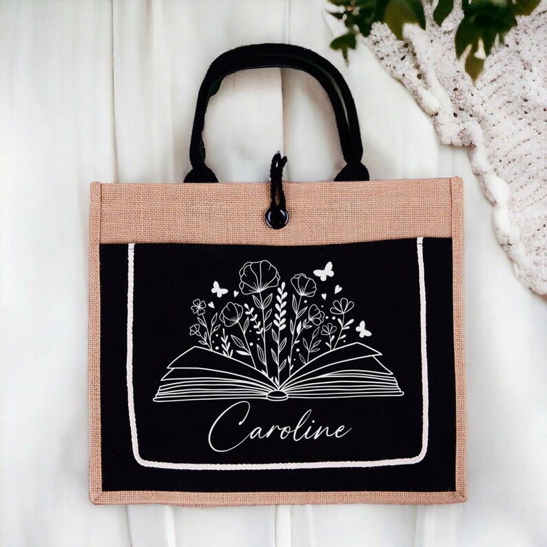 May include: A black and beige tote bag with a white design of an open book with flowers and butterflies. The name "Caroline" is written below the book.