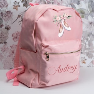 May include: A light pink backpack with a front pocket and a gold zipper. The backpack features a watercolor design of ballet shoes and the name "Audrey" in a cursive font. The backpack has pink straps.