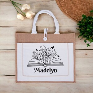 May include: A white tote bag with a brown burlap exterior. The bag has a white pocket with a black and white illustration of an open book with flowers and butterflies. The text "Madelyn" is printed below the illustration.