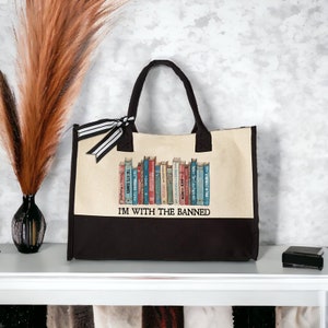 Peut inclure: Un sac fourre-tout en toile noir et blanc avec un panneau en toile beige repr&eacute;sentant une rang&eacute;e de livres color&eacute;s avec le texte "I'M WITH THE BANNED".