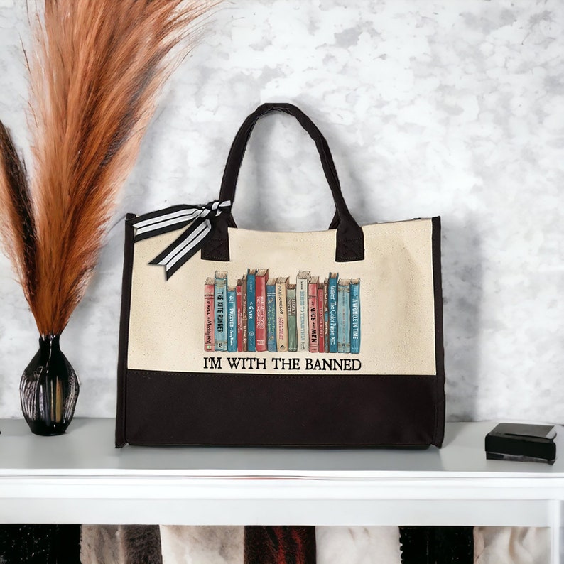 Peut inclure: Un sac fourre-tout en toile noir et blanc avec un panneau en toile beige repr&eacute;sentant une rang&eacute;e de livres color&eacute;s avec le texte "I'M WITH THE BANNED".