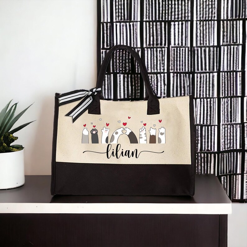May include: A black and beige tote bag with a design of cats holding paws and hearts. The bag has a black handle and a white stripe on the handle. The name "lilian" is printed on the bag.