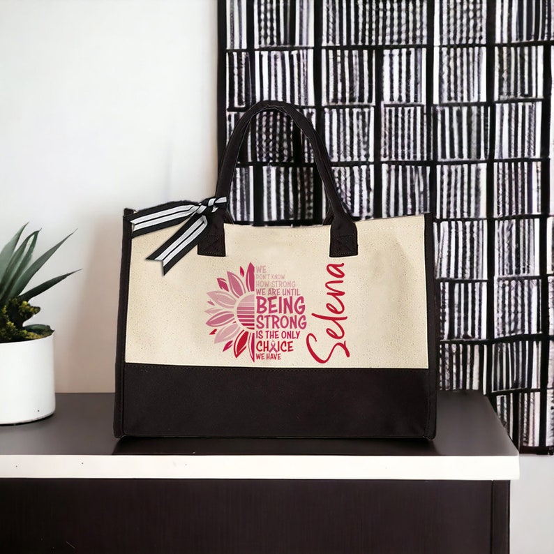 May include: A beige and black canvas tote bag with a sunflower design and the text "WE DON'T KNOW HOW STRONG WE ARE UNTIL BEING STRONG IS THE ONLY CHOICE WE HAVE". The name "Selena" is written vertically on the bag.