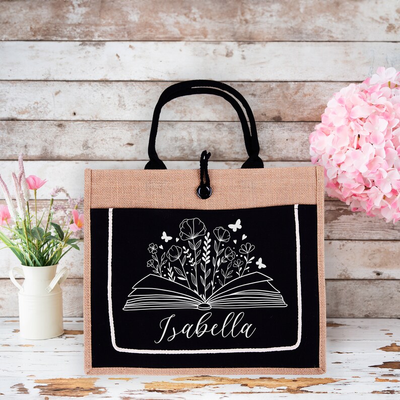 May include: A brown and black tote bag with a black fabric pocket featuring a white outline of an open book with flowers and butterflies. The name "Isabella" is written in white below the book.