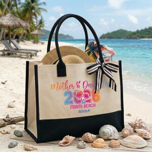 Pu&ograve; includere: Una borsa tote in tela beige con manici neri e fondo nero. La borsa presenta la scritta "Mother & Daughter 2026 Miami Beach anne" in lettere colorate. Un cappello di paglia e una sciarpa fantasia sono all'interno.