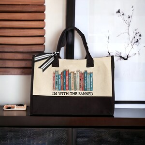 Peut inclure: Un sac fourre-tout en toile noire avec un panneau en toile beige repr&eacute;sentant une rang&eacute;e de livres illustr&eacute;s. Le panneau beige porte l'inscription "I'M WITH THE BANNED".
