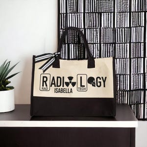 May include: A black and beige canvas tote bag with the words "RADIOLOGY" in black lettering. The bag has a black skull graphic and the words "RAD", "TECH", and "ISABELLA" in white lettering.