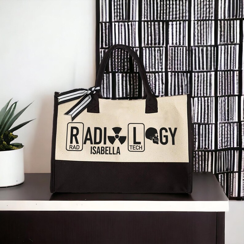 May include: A black and beige canvas tote bag with the words "RADIOLOGY" in black lettering. The bag has a black skull graphic and the words "RAD", "TECH", and "ISABELLA" in white lettering.