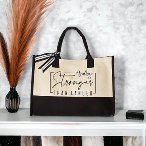 May include: A beige and black canvas tote bag with black handles and a black and white striped ribbon. The bag has the text "Audrey Stronger Than Cancer" printed on the front.