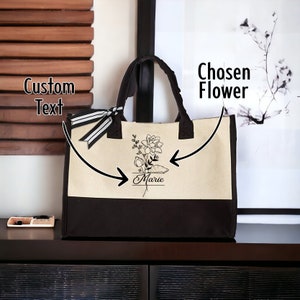 May include: A black and cream tote bag with a black and white ribbon. The bag has a black floral design with the name "Marie" written underneath. The text "Chosen Flower" is written on the right side of the image and the text "Custom Text" is written on the left side of the image.