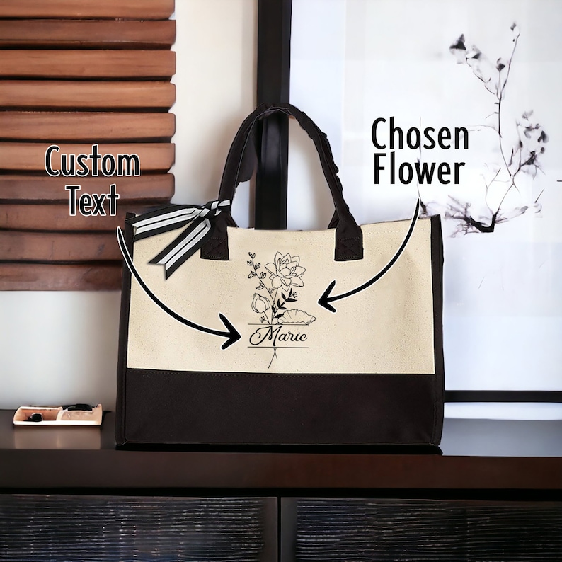 May include: A black and cream tote bag with a black and white ribbon. The bag has a black floral design with the name "Marie" written underneath. The text "Chosen Flower" is written on the right side of the image and the text "Custom Text" is written on the left side of the image.