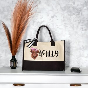 K&ouml;nnte beinhalten: Eine braune und wei&szlig;e Tragetasche mit schwarzem Boden. Die Tasche hat ein schwarz-wei&szlig;es Band und eine Comic-Highland-Kuh mit einer rosa Schleife auf dem Kopf. Die Tasche ist mit dem Namen "Ashley" personalisiert.