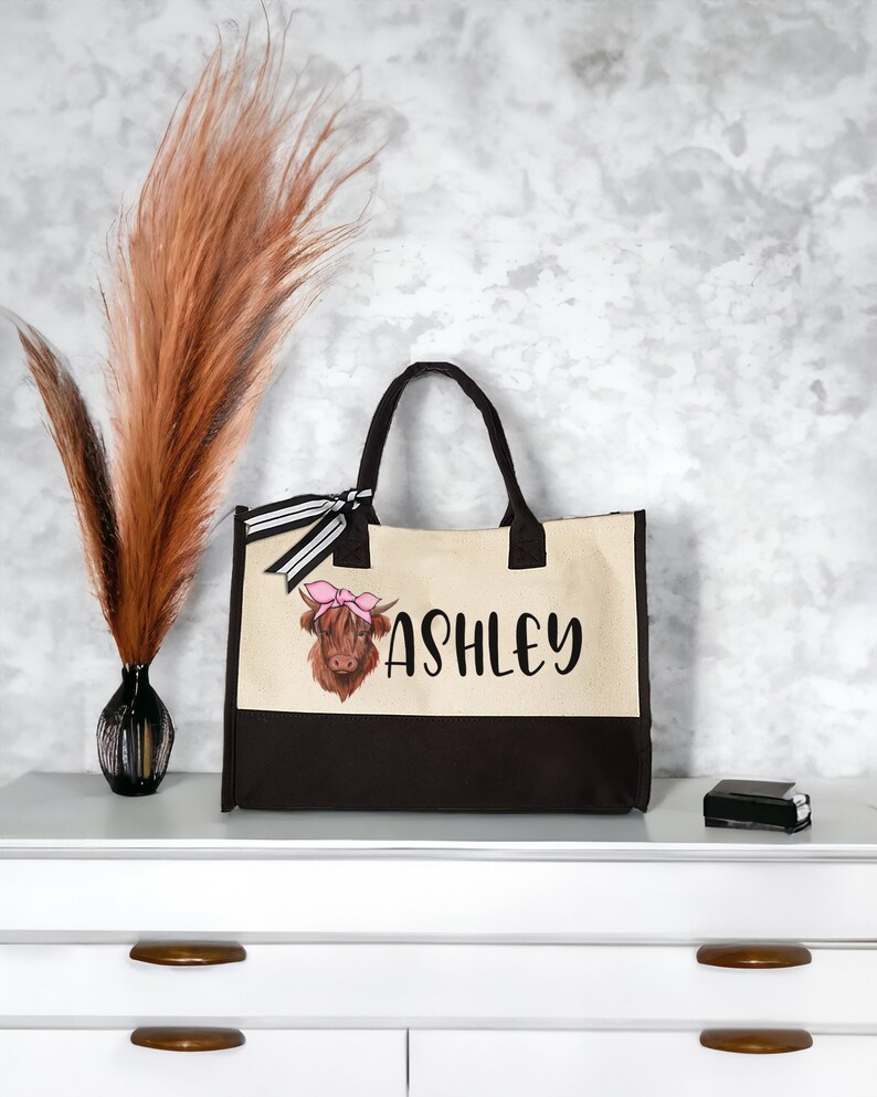 K&ouml;nnte beinhalten: Eine braune und wei&szlig;e Tragetasche mit schwarzem Boden. Die Tasche hat ein schwarz-wei&szlig;es Band und eine Comic-Highland-Kuh mit einer rosa Schleife auf dem Kopf. Die Tasche ist mit dem Namen "Ashley" personalisiert.