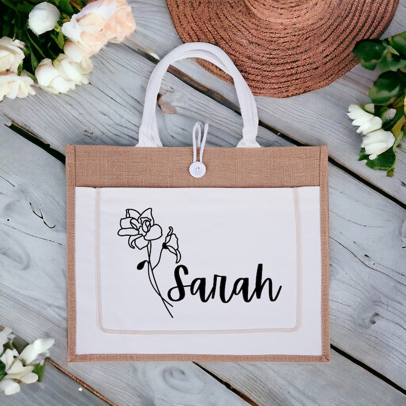 May include: A brown and white tote bag with a white pocket featuring a black line drawing of a flower and the name "Sarah".