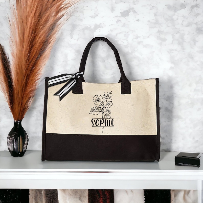 May include: A beige and black canvas tote bag with black and white striped ribbon handles. The bag features a floral design and the name "Sophie" printed on the front.
