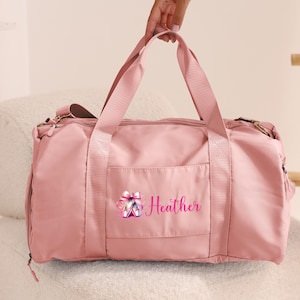 May include: Pink duffel bag with a zipper closure and a front pocket. The bag has a pink ballerina shoe design and the name "Heather" embroidered on the front.