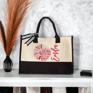 May include: A beige and black canvas tote bag with a pink and white sunflower design. The bag has a black and white striped ribbon and the text "WE DON'T KNOW HOW STRONG WE ARE UNTIL BEING STRONG IS THE ONLY CHOICE WE HAVE" and the name "Emilia" printed on it.