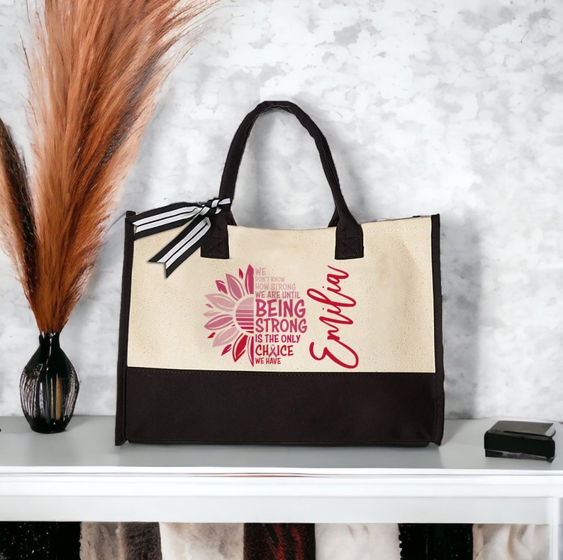 May include: A beige and black canvas tote bag with a pink and white sunflower design. The bag has a black and white striped ribbon and the text "WE DON'T KNOW HOW STRONG WE ARE UNTIL BEING STRONG IS THE ONLY CHOICE WE HAVE" and the name "Emilia" printed on it.