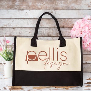 May include: A beige and black canvas tote bag with the text "pellis design" printed in brown on the front. The bag has two black handles.