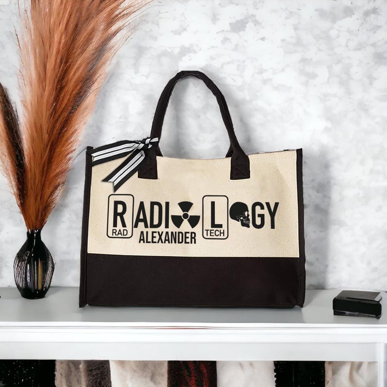 May include: A beige and black canvas tote bag with the words "RADIOLOGY" in black lettering. The word "RAD" is in a smaller box above the word "ALEXANDER" which is in a larger box. The word "TECH" is in a smaller box above the word "LOGY" which is in a larger box. A black and white radiation symbol is in the box with the word "ALEXANDER". A black and white skull is in the box with the word "LOGY".