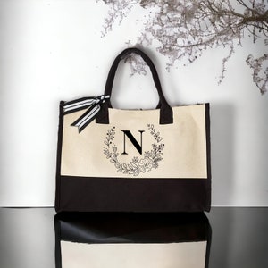 May include: A beige and brown canvas tote bag with a black floral wreath and the letter "N" in the center.