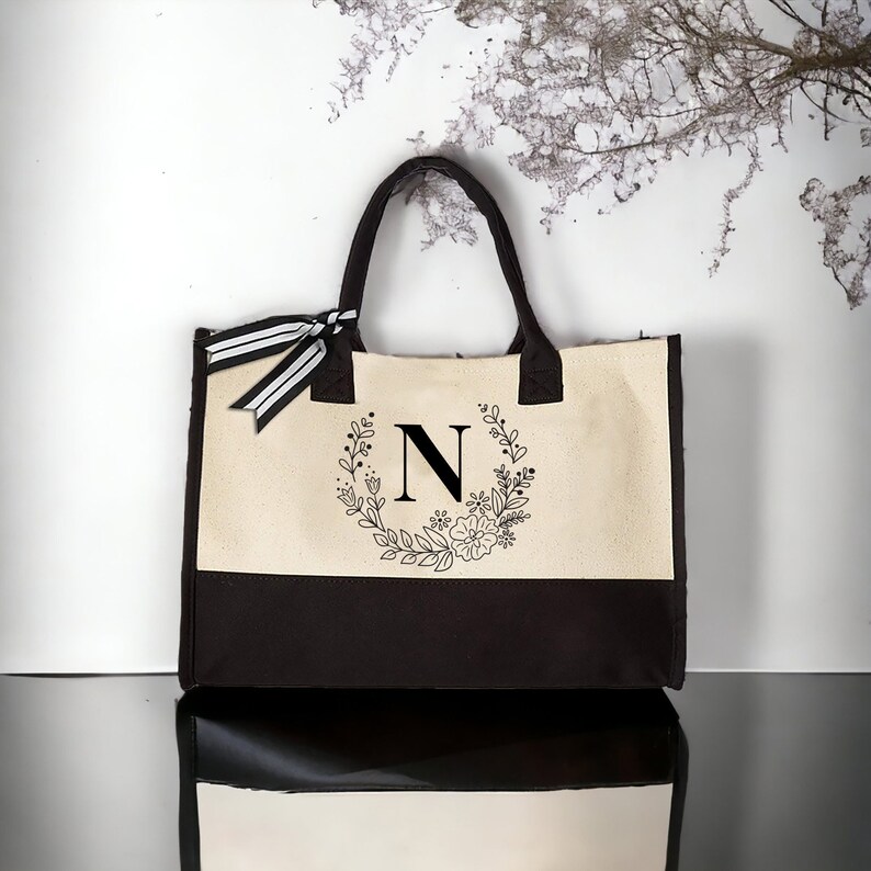 May include: A beige and brown canvas tote bag with a black floral wreath and the letter "N" in the center.