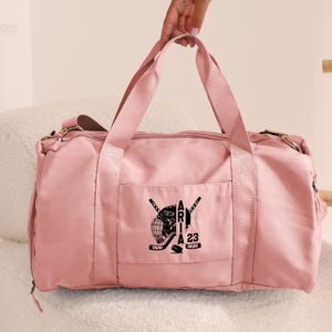 May include: A pink duffel bag with a black hockey-themed design featuring a hockey helmet, crossed hockey sticks, and the text "Aria 23".