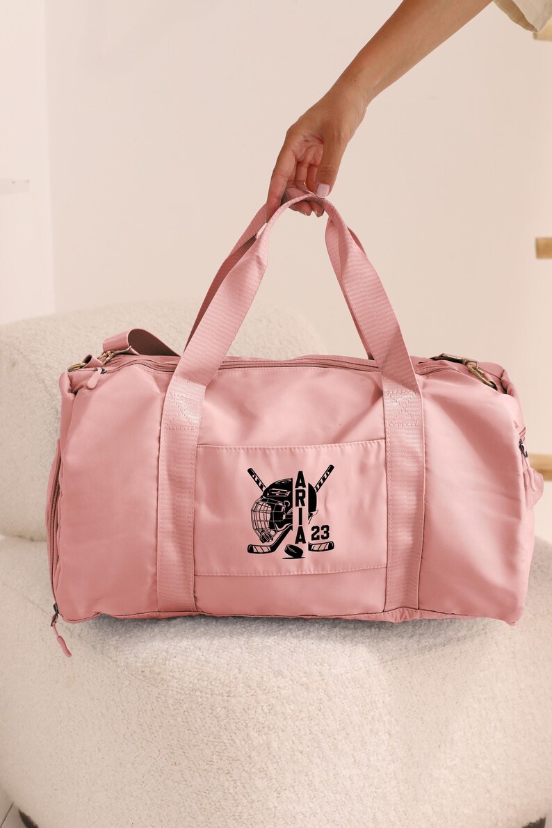May include: A pink duffel bag with a black hockey-themed design featuring a hockey helmet, crossed hockey sticks, and the text "Aria 23".