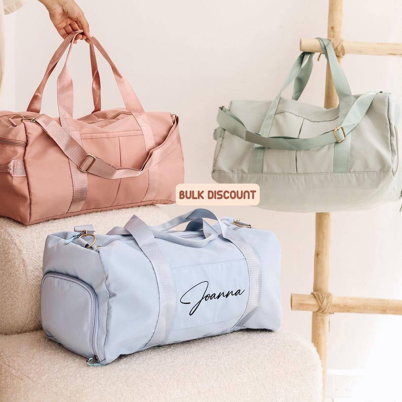 May include: Three duffel bags in pastel colors: pink, mint green, and light blue. The light blue bag has the name "Joanna" printed on it. The bags have shoulder straps and side pockets. The text "BULK DISCOUNT" is visible.