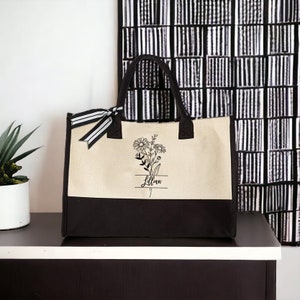 May include: A beige and black canvas tote bag with a floral design and the name "Lillian" printed on it.