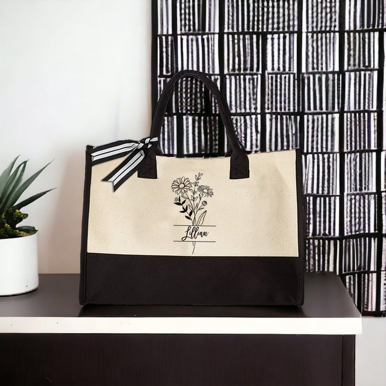 May include: A beige and black canvas tote bag with a floral design and the name "Lillian" printed on it.