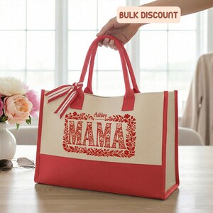 May include: A canvas tote bag with red accents and handles. The bag features the word "MAMA" in a floral design, with the name "Ashley" above it. A red and white striped ribbon is tied to the handle. The bottom of the bag is red.