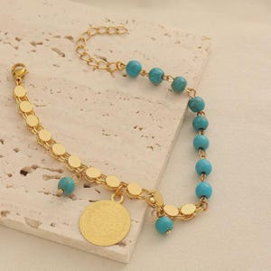 Puede incluir: Pulsera dorada con una cadena de pequeños círculos planos y cuentas turquesas. Un colgante dorado con un diseño grabado cuelga de la pulsera. La pulsera tiene un cierre de mosquetón.