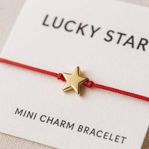 Puede incluir: Una pulsera con dije de estrella dorada con un cordón rojo. La pulsera se muestra en una tarjeta blanca con las palabras "LUCKY STAR" y "MINI CHARM BRACELET" impresas en negro.