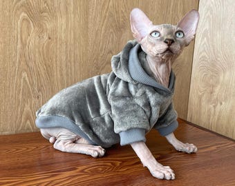 Puede incluir: Un suéter gris con capucha para un gato. El suéter está hecho de tela suave y afelpada y tiene una capucha con un borde acanalado. El gato es de raza Sphynx y tiene un pelaje gris claro.