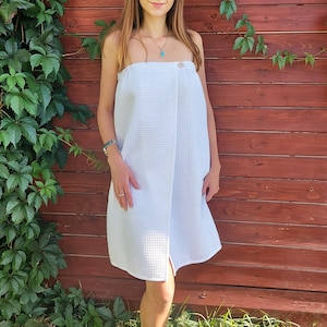 May include: A white waffle weave wrap with a button closure. The wrap is worn by a person standing in front of a wooden wall with vines.