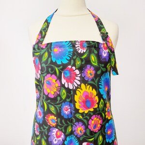 May include: A black apron with a colorful floral pattern. The apron has a halter neck style and is made of a cotton fabric.