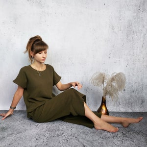 May include: A woman wearing a dark green, loose-fitting, short-sleeved top and matching pants sits on a light gray textured surface. She is looking down at her hands, which are resting on her lap. There is a dried flower arrangement in a brown vase to the right of the woman.