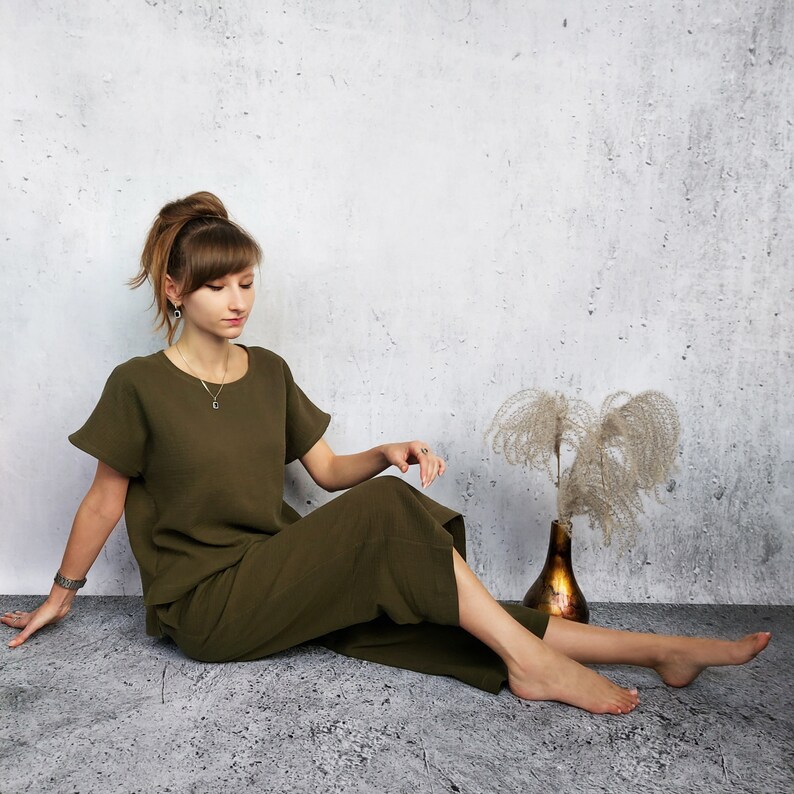 May include: A woman wearing a dark green, loose-fitting, short-sleeved top and matching pants sits on a light gray textured surface. She is looking down at her hands, which are resting on her lap. There is a dried flower arrangement in a brown vase to the right of the woman.