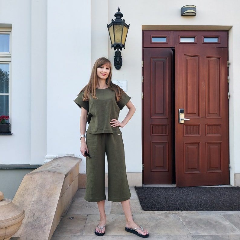 May include: A woman wearing a green two-piece outfit with a short-sleeved top and cropped pants. She is standing in front of a wooden door with a light fixture above.